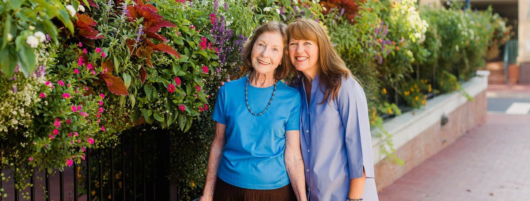 elderly woman and adult daughter smiling in sunshine near wall of flowers